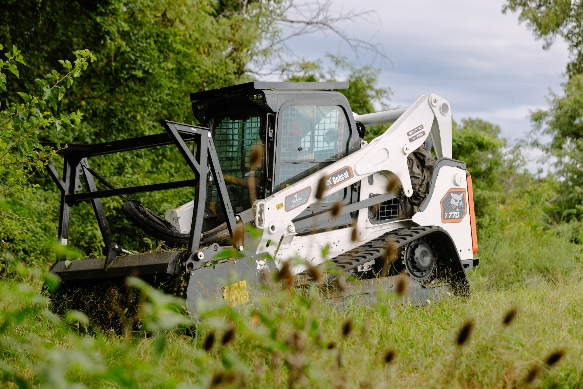Skid steer with brush hog attachment cutting heavy growth across an overgrown pasture