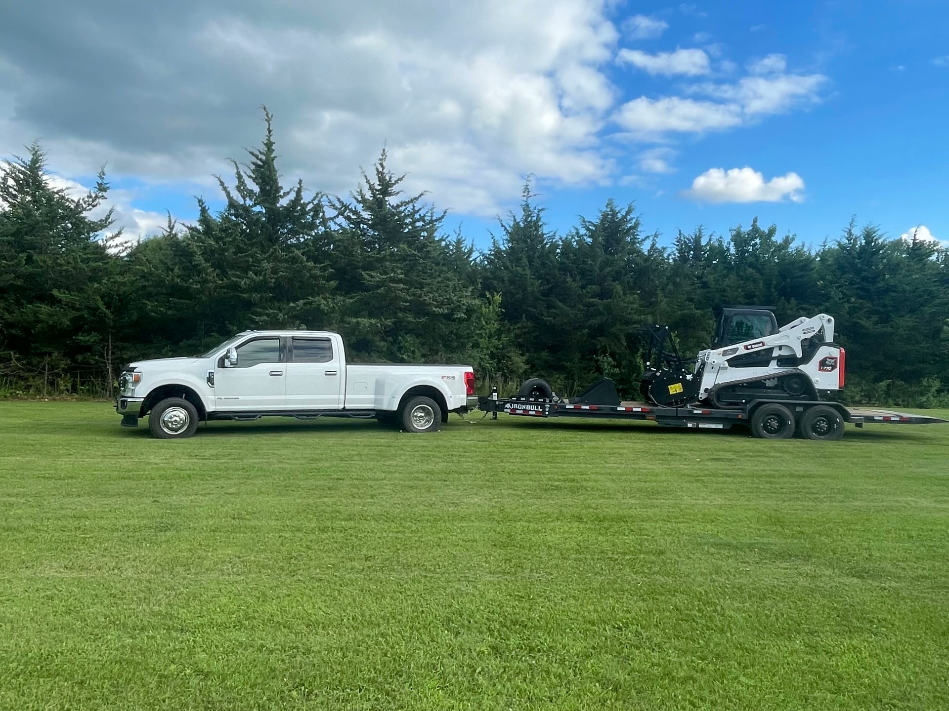 Work truck and equipment trailer carrying a compact track loader and forestry mulcher, parked on a grass lawn